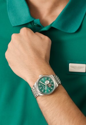 Wrist with stainless steel watch featuring green dial, open-heart design, and minute markers, worn over green collared shirt with badge.