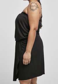 Nainen, jolla on letitetty hiustyyli ja olkaimeton musta mekko, esittelee yläkädessään pyöreää tatuointia ja ranteessaan herkän rannekorun.