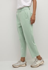 Pantalon ajusté vert menthe avec plis à l'avant, poches latérales et longueur cropped, associé à des baskets gris clair avec une semelle texturée.