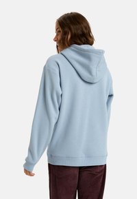 Sudadera con capucha de color azul claro, con cordones, textura suave y puños acanalados. Llevada con pantalones de pana oscuros, que muestran un ajuste relajado.