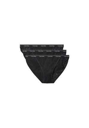 Femme aux longs cheveux bruns portant un top court noir et un sous-vêtement noir Calvin Klein avec un détail en dentelle, debout devant un fond gris uni.