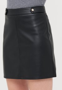 Mini jupe en cuir noir avec une texture lisse, présentant une coupe ajustée et deux boutons métalliques décoratifs à la taille.