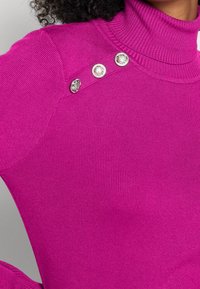 Gros plan sur une personne portant un pull à col roulé côtelé rose vif avec trois boutons décoratifs argentés sur l'épaule.