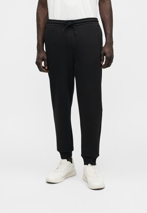 Άνδρας που φοράει μαύρο παντελόνι τζόγκινγκ με μανσέτες στους αστραγάλους, λευκό μπλουζάκι και λευκά αθλητικά παπούτσια, στέκεται μπροστά σε ανοιχτό γκρι φόντο.