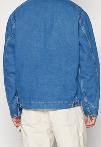 Modrá džínová bunda s uvolněným střihem, zdobená zlatými prvky, s rovním lemem a kontrastním prošitím. Hladká textura a ležérní design.