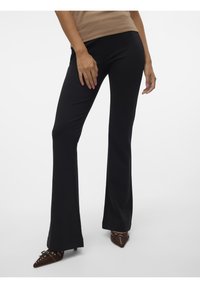 Pantalon noir évasé en tissu doux, caractérisé par une texture lisse. Associé à des talons bruns pointus et un haut beige ajusté.