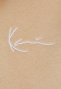 Tessuto lavorato a maglia beige con una texture liscia, caratterizzato da testo ricamato bianco che dice "Kani", posizionato al centro.