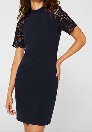 Femme portant une robe bleu marine cintrée avec des manches courtes en dentelle et un col montant, posant avec une main sur la hanche.