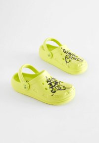 Neon sárga klumpák sima műanyag felülettel, szellőzőlyukakkal és fekete graffitis feliratokkal. Hátsó pánt a biztonságos illeszkedés érdekében.
