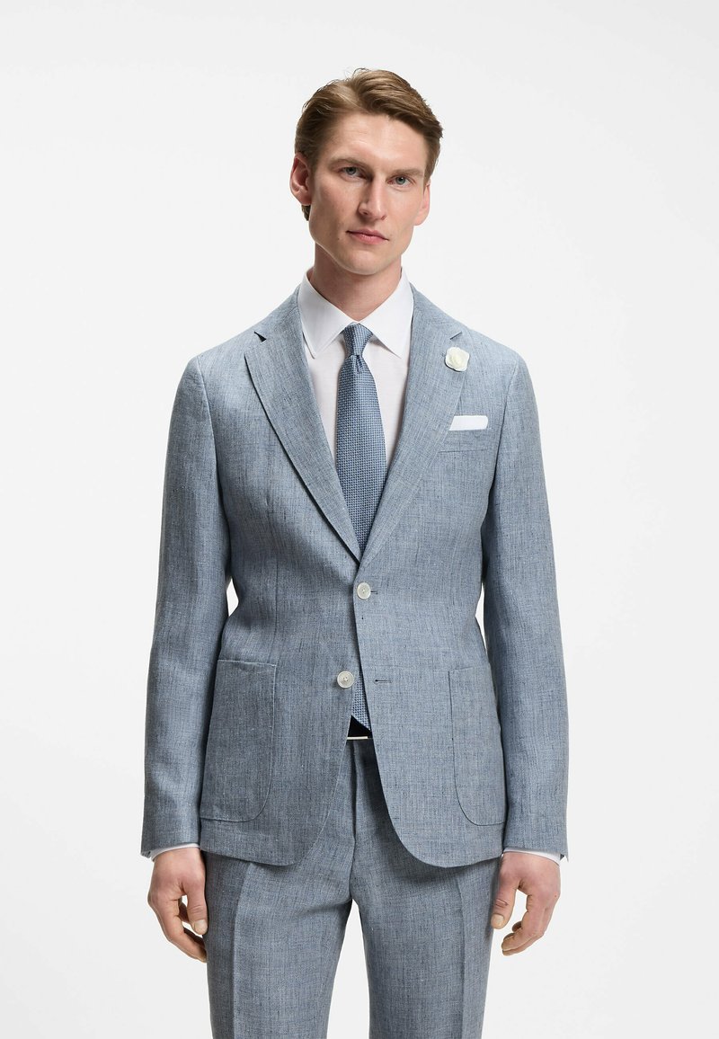 Homme portant un costume bleu clair, une chemise blanche, une cravate bleue texturée et une épingle de revers blanche, debout devant un fond blanc uni.