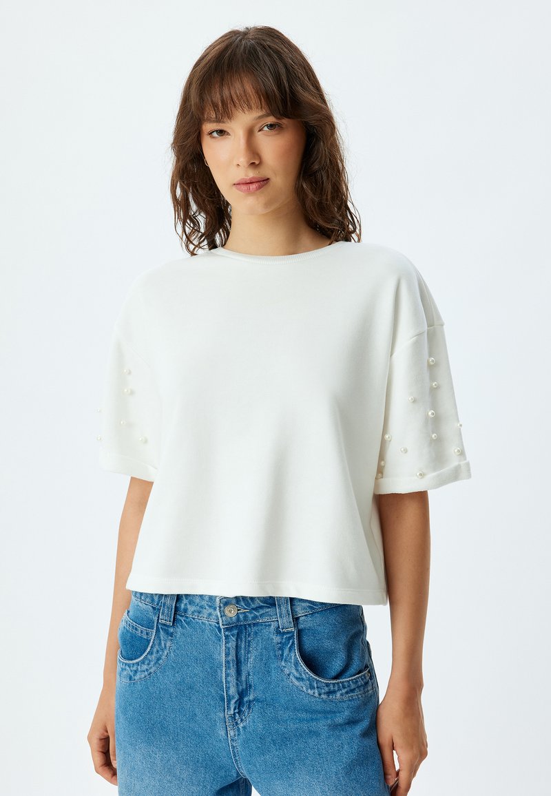 Valkoinen lyhythihainen crop-top, jossa on koristeelliset helmikoristeet hihoissa. Yhdistetty sinisiin farkkuihin.