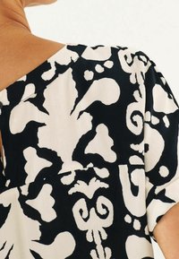 Schwarz-weiße gemusterte Bluse mit fließendem Design, weicher Textur und einem Rundhalsausschnitt mit asymmetrischem Schnitt.