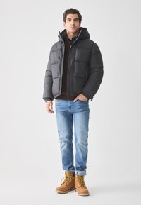Hombre joven vistiendo una chaqueta de plumas negra, un suéter marrón, jeans azul claro y botas de trabajo color beige, de pie contra un fondo blanco liso.