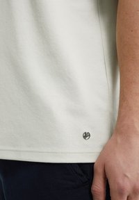 Soehker grey puuvillane T-särk, millel on sile tekstuur ja väike metallist logo aktsent allääril. Lühikeste varrukatega disain on nähtav.