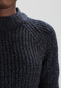 Marineblauer Strickpullover mit hohem Kragen, der dicke, strukturierte Garne und subtile metallische Fäden im gesamten Stoff aufweist.