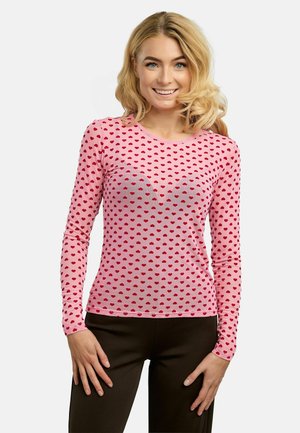 Femeie blondă zâmbitoare, purtând o cămașă roz cu mâneci lungi, cu modele de inimioare roșii și pantaloni negri, stând în fața unui fundal deschis, simplu.