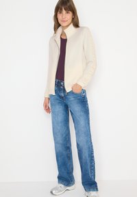 Chaqueta de cremallera en beige claro con un cuello alto, combinada con una blusa morada oscura y jeans de pierna ancha azules. El modelo está de pie con las manos en los bolsillos.