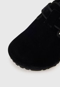 Chaussure slip-on en suede noir avec un bout arrondi, présentant un détail de sangle et une semelle texturée pour une meilleure adhérence. Design minimaliste avec une finition lisse.