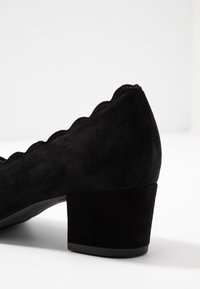 Chaussure à talon en daim noir avec un design à bords festonnés, dotée d'un talon carré et d'une texture lisse. Gros plan sur la région du talon.