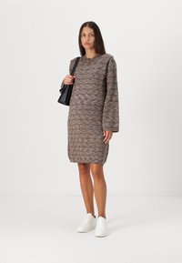 Robe en tricot à manches longues dans des tons marron et gris, avec une coupe décontractée et un ourlet côtelé. Associée à des baskets blanches et un sac noir.