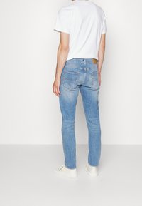 Una persona se encuentra de espaldas al espectador, vistiendo una camiseta blanca y unos jeans azul claros, combinados con zapatos blancos frente a un fondo neutral.