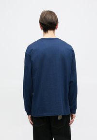 Henkilö nähtynä takaa, päällä sininen pitkähihaisuus paita ja mustat housut, lyhyet ruskeat hiukset ja pienet renkaat korvissa.