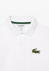 Polo blanc en tissu respirant, avec un col vert, trois boutons et un logo crocodile brodé en vert et rouge sur la poitrine.