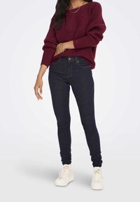 Maglione lavorato a maglia color mogano con maniche ampie abbinato a jeans skinny blu scuro. Il modello indossa scarpe da ginnastica bianche; la posizione rilassata seduta esalta il look casual.