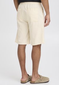 Shorts beige clair avec une ceinture élastique et deux poches arrière, présentant un motif texturé subtil. Portés avec des chaussures grises à enfiler.