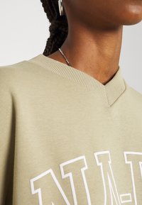 Sudadera verde claro con un diseño de escote en V, cuello de canalé y letras gráficas blancas. El material parece suave y texturizado.