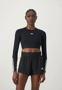 Fekete hosszú ujjú crop top fehér logóval, fekete, magas derekú rövidnadrággal párosítva, fehér oldalsávokkal. Sportos dizájn és illeszkedés.