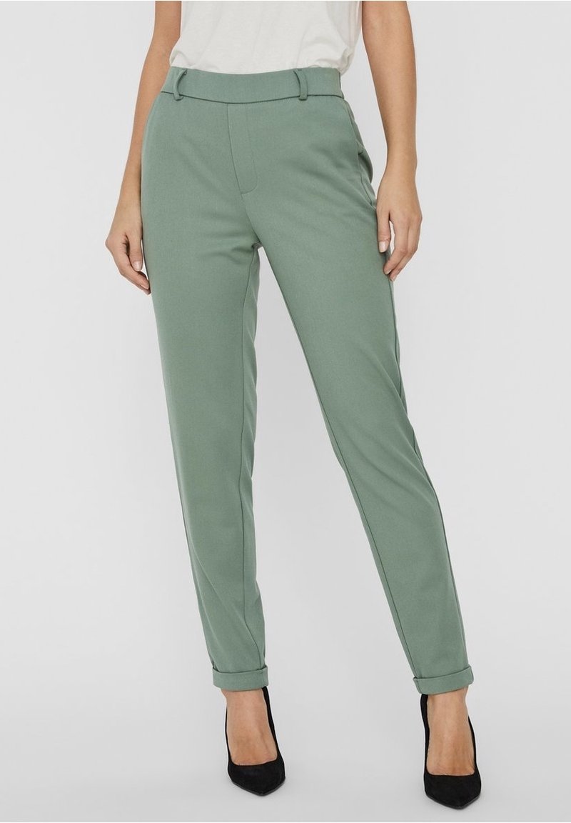 Femme portant un pantalon vert sauge cintré avec ourlets retroussés et des escarpins noirs à bout pointu, associé à un haut blanc, debout sur un sol gris.