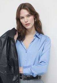 Camicia button-up in satin blu chiaro con colletto, indossata sotto una giacca di pelle nera. Modello in posa con una vita nera cinturata. Texture liscia, design casual.