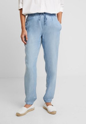 Person trägt hellblaue Kordelhose mit elastischen Bündchen und weiße Espadrilles zum Hineinschlüpfen und steht vor schlichtem Hintergrund.