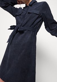 Abito camicia in corduroy blu navy con maniche lunghe, tasche frontali e cintura in vita. Presenta una texture a coste verticali e un design con bottoni.