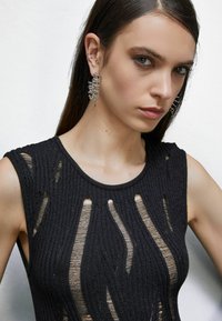 Top nero in maglia con texture a costine e dettagli a taglio; presenta un collo ampio e una design delle spalle. Accessoriato con grandi orecchini in argento.