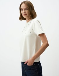 Weißes T-Shirt mit kurzen Ärmeln, rundem Halsausschnitt und verstreuten dekorativen Nieten auf der Vorderseite, kombiniert mit dunkelblauen Jeans.
