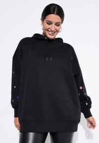 Černá oversized mikina vyrobená z měkké látky, se zlatými kamínkovými ozdobami na obou rukávech a stahovací kapucí.