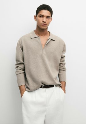 Junger Mann trägt einen beigen langärmligen Polo-Pullover und weiße Hose mit einem dunkel geflochtenen Gürtel, steht mit den Händen in den Taschen.