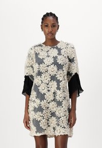 Kort kjole laget av gjennomsiktig stoff med et beige blomsterdesign over en grå prikkete bakgrunn, med svarte plisserte ermer.