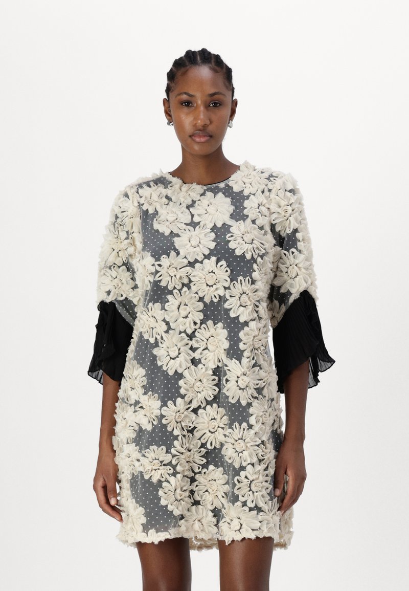 Kort kjole laget av gjennomsiktig stoff med et beige blomsterdesign over en grå prikkete bakgrunn, med svarte plisserte ermer.