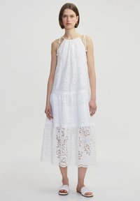 Vestido blanco sin mangas con patrones florales bordados, escote fruncido y encaje en el dobladillo. Modelo lleva sandalias planas blancas.