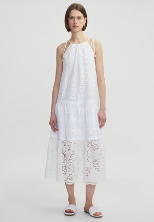 Vestido blanco sin mangas con patrones florales bordados, escote fruncido y encaje en el dobladillo. Modelo lleva sandalias planas blancas.