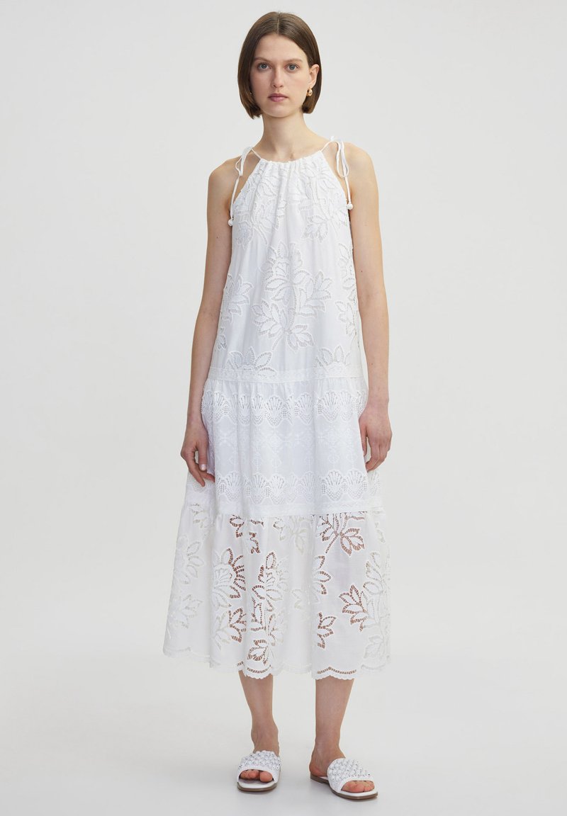 Vestido blanco sin mangas con patrones florales bordados, escote fruncido y encaje en el dobladillo. Modelo lleva sandalias planas blancas.