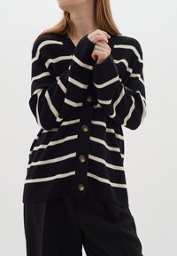 Námořnický kardigan s bílými pruhy, s výstřihem do V, zapínáním na knoflíky a dlouhými rukávy. Měkká pletená textura s dvěma bočními kapsami.