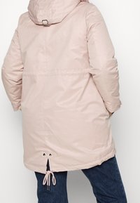 Person in einem langen, hellrosa gepolsterten Mantel mit Kapuze, der am Saum einen Kordelzug hat, kombiniert mit dunklen Jeans.