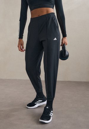 Femme en pantalon de sport noir, haut court à manches longues et baskets noires tenant une kettlebell dans une main.