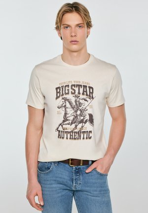 Młody mężczyzna o blond włosach, ubrany w beżowy t-shirt z nadrukiem jeźdźca konnego i niebieskie dżinsy, stojący z ręką w kieszeni.