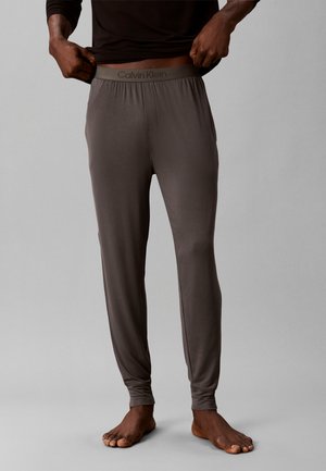 Homme portant un pantalon de sous-vêtements longs gris Calvin Klein, remontant un t-shirt noir, debout pieds nus sur un fond gris.