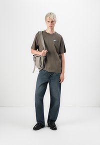 Grijze katoenen t-shirt met logo, gecombineerd met een slouchy paar donkerblauwe denim jeans. Model draagt een kleine grijze tas met ritsdetails. Zwarte schoenen.